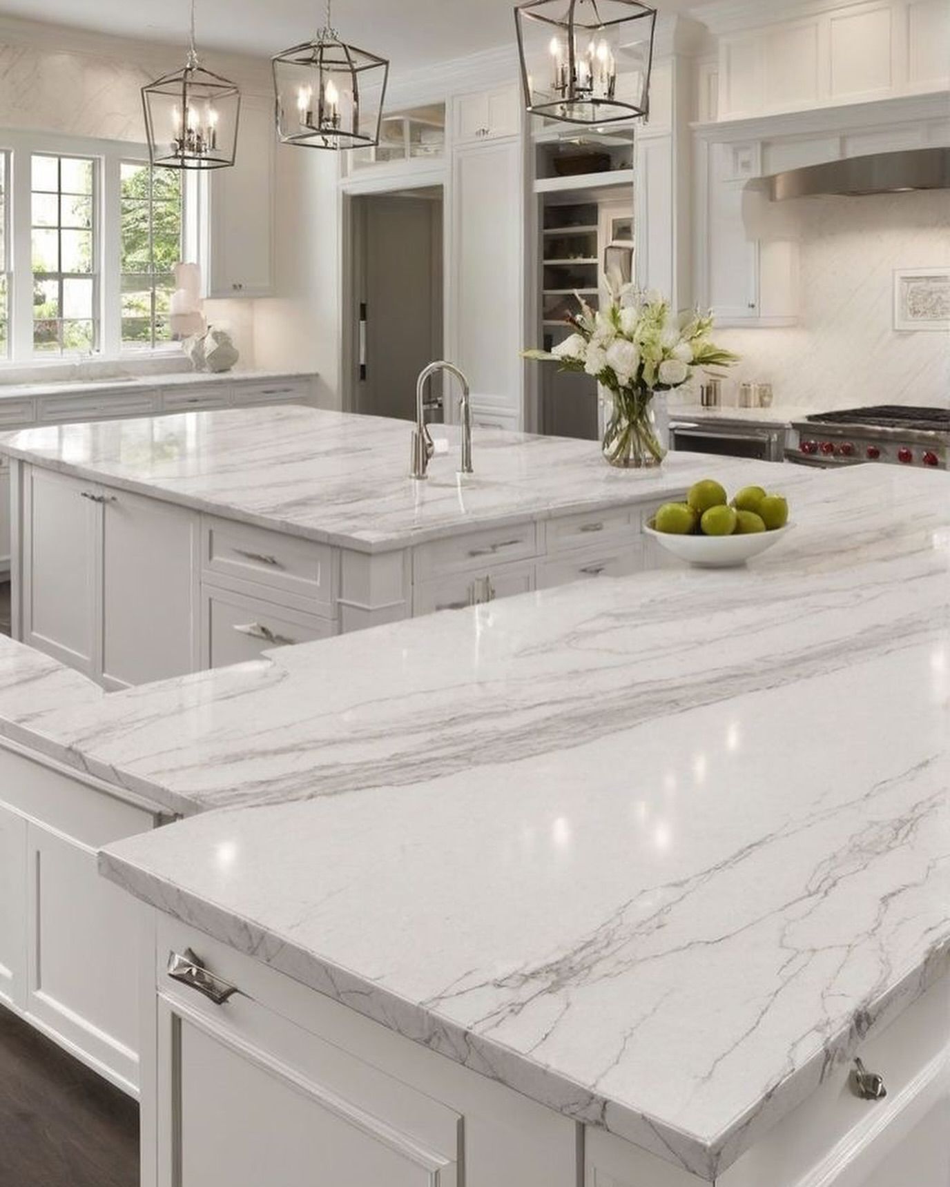 Bright, white kitchen with two marble-topped islands and hanging lights. Features a stove, cabinets, and a vase of flowers.