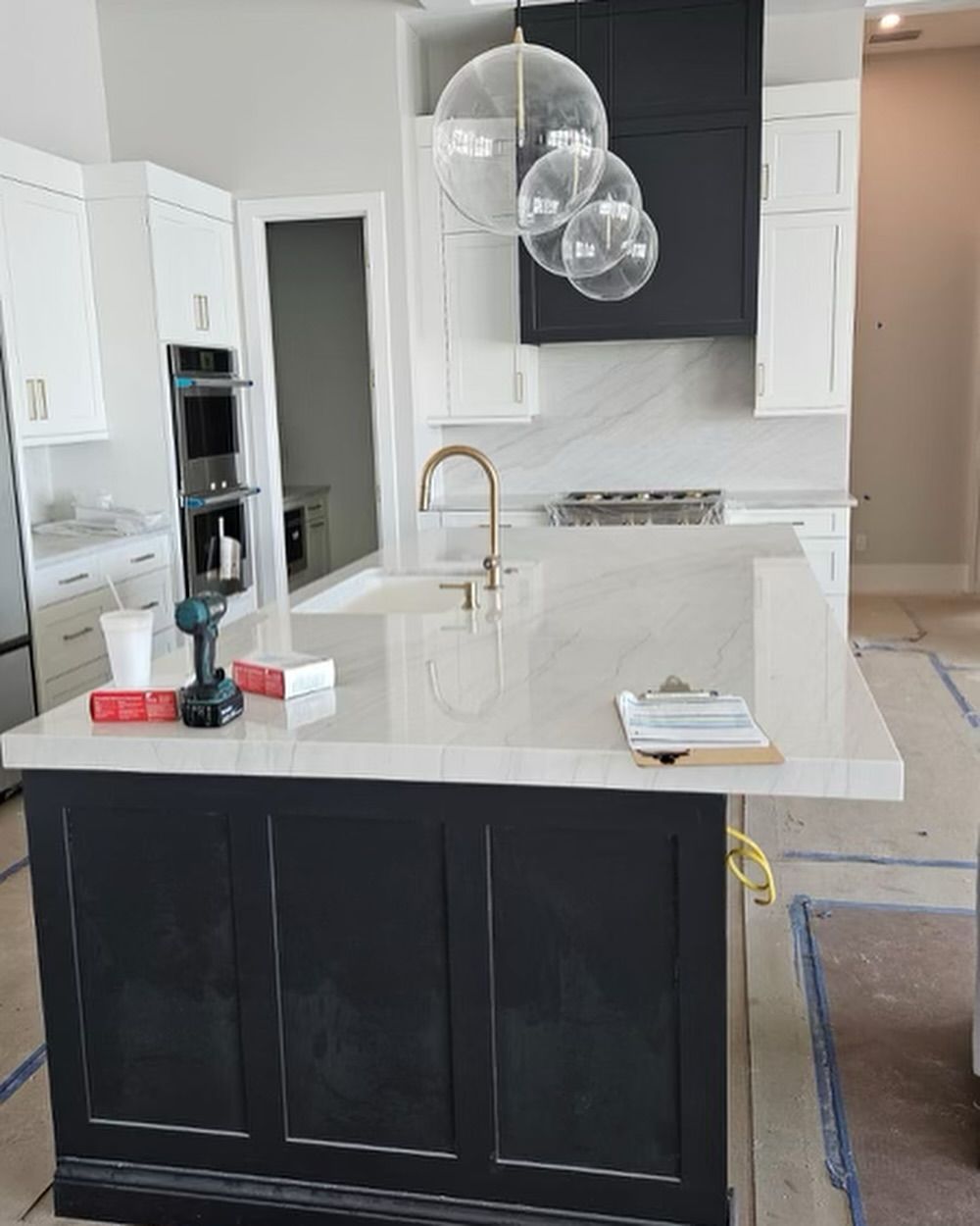 Kitchen with black island, white cabinets, and gold faucet, unfinished floor, clear bubble pendant lights.