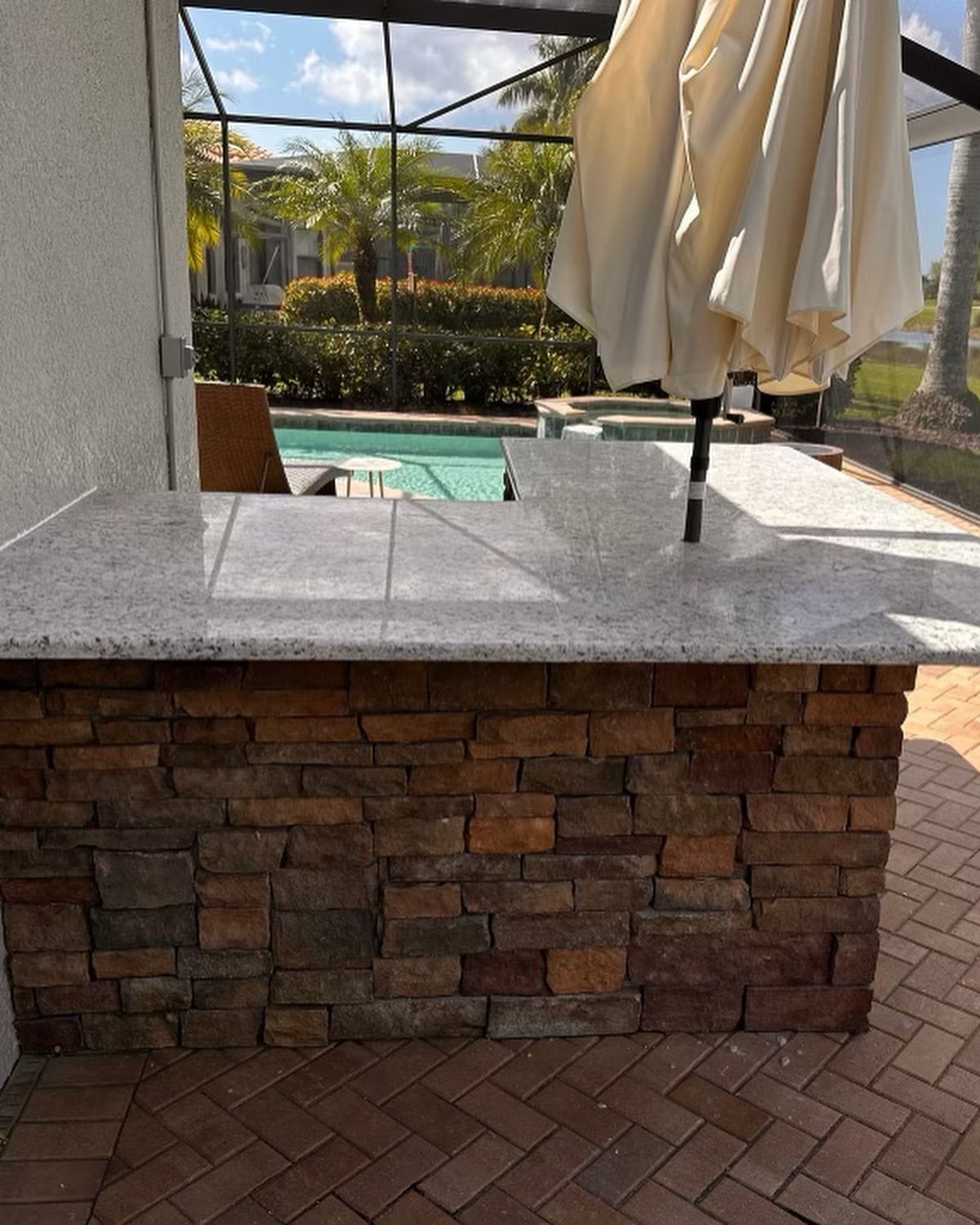 Outdoor bar with stone facade and light-colored countertop, next to a pool under a screened enclosure.
