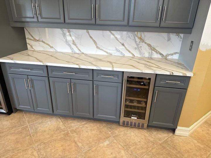 Gray cabinets with white marble countertop and backsplash, built-in wine fridge, tile floor.