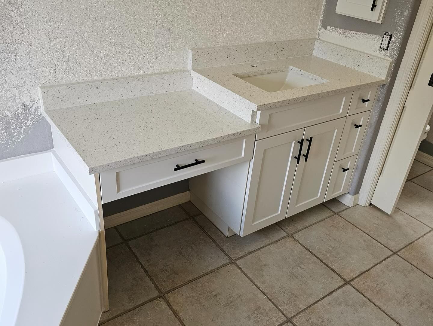 White bathroom vanity with quartz countertop, sink, cabinets, and drawer, against a tiled wall.