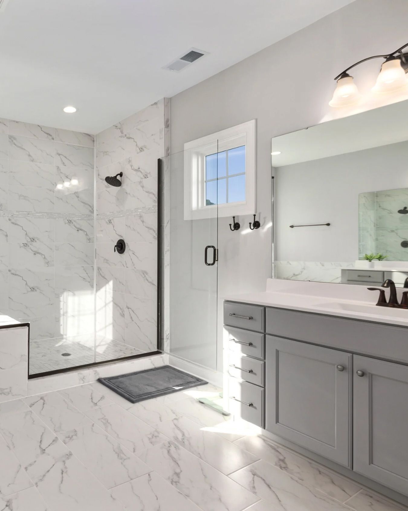Spacious, modern bathroom with marble tile and gray vanity. Walk-in shower with black fixtures and window.