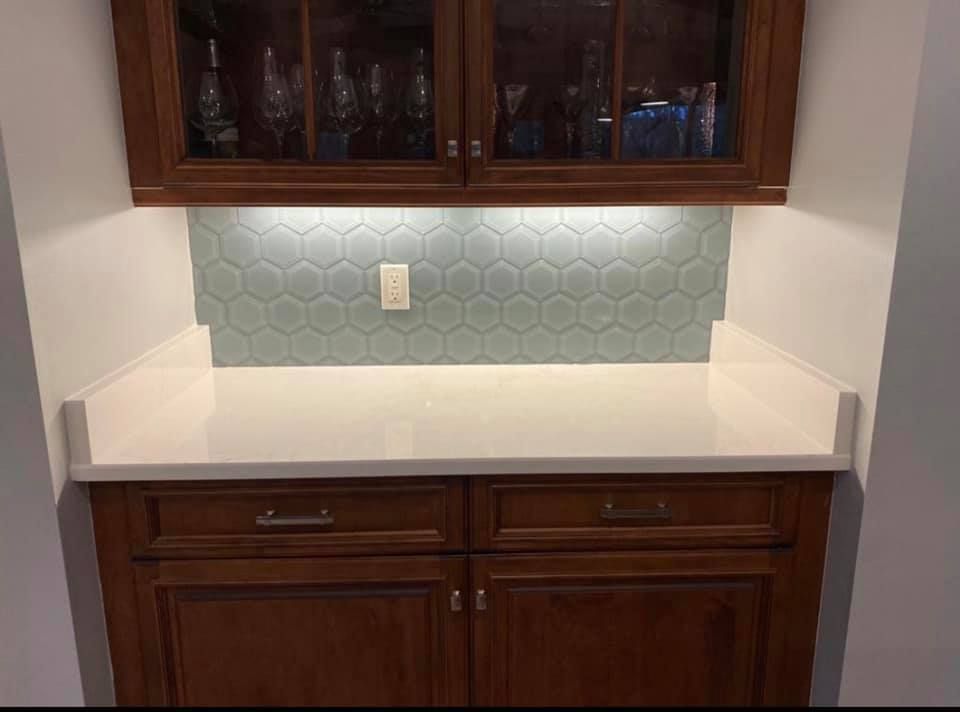 Built-in bar with wooden cabinets, white countertop, blue hexagon tile backsplash, and overhead lighting.