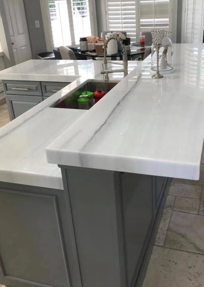 Gray kitchen island with white countertop and sink, looking into another room with blinds.