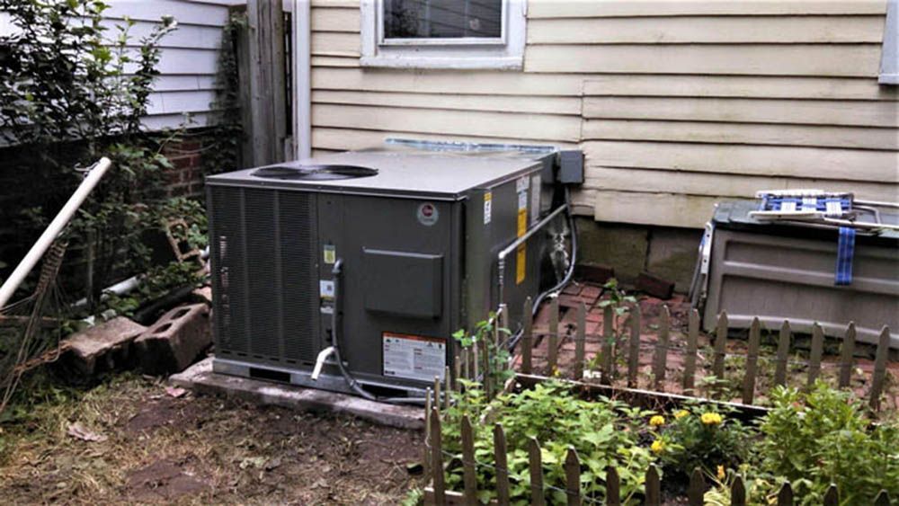 Large gray HVAC unit beside a beige house, with a small garden and fence in the foreground.