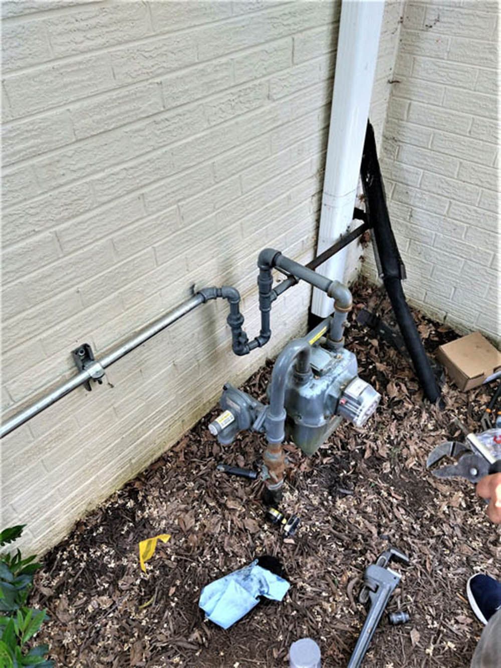 Gas meters and utility pipes mounted on an exterior beige brick wall near mulch and debris.