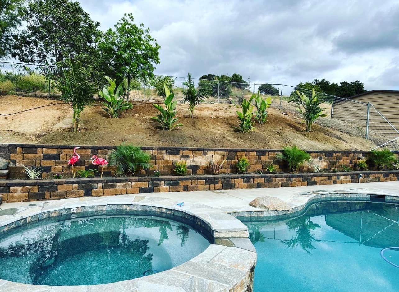 Pool and spa overlook a retaining wall with landscaping and a sloped hillside under an overcast sky.