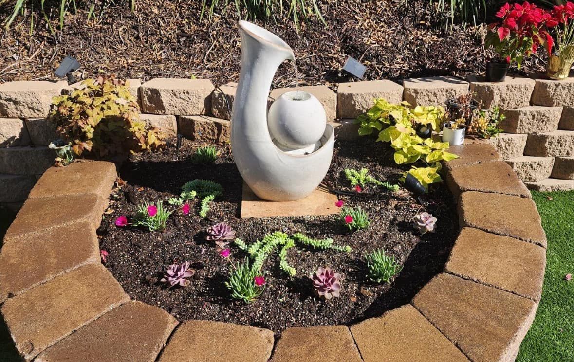 Stone fountain in a circular garden bed, surrounded by a brick border, with flowers and plants.