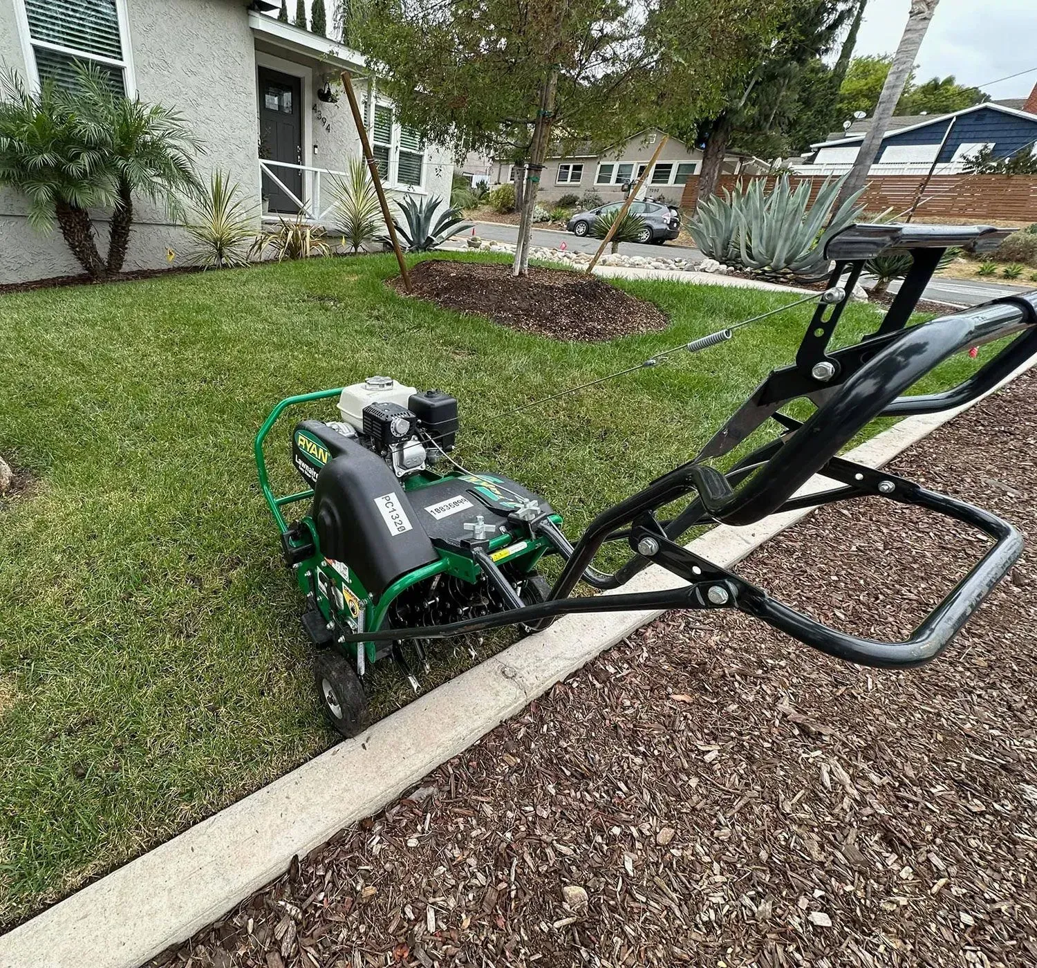 Green lawn aerator on a lawn, next to a brown mulch bed. Green grass, house in background.