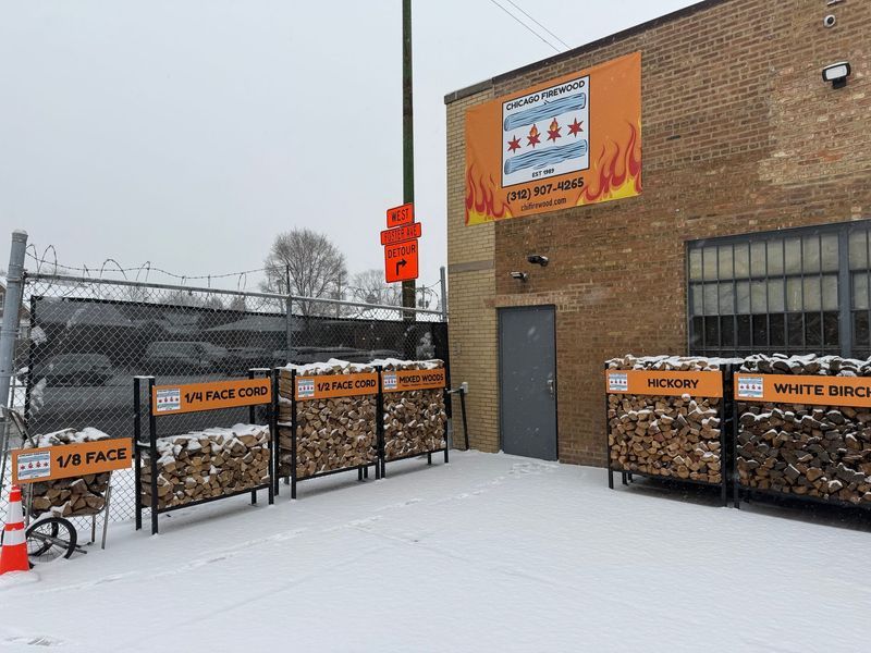 A snowy scene with a firewood vendor in Chicago. Firewood in labeled bins and an orange sign with the Chicago flag.