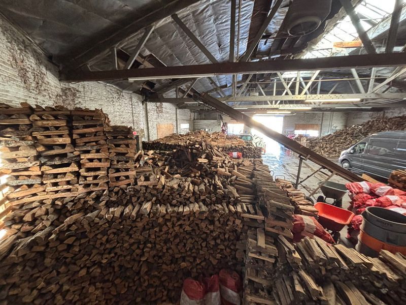 Warehouse filled with stacked firewood and pallets, with sunlight streaming in.