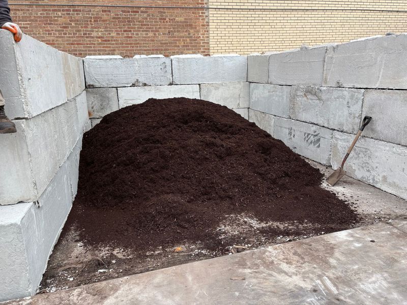 Pile of dark mulch within a concrete block enclosure, with a shovel resting nearby.