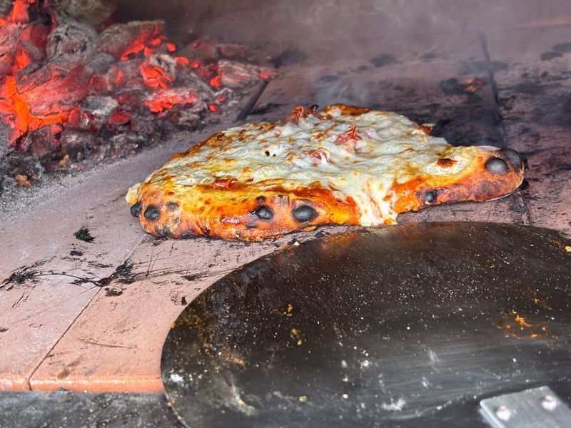Pizza baking in a wood-fired oven with melted cheese and a charred crust; a pizza peel rests below.