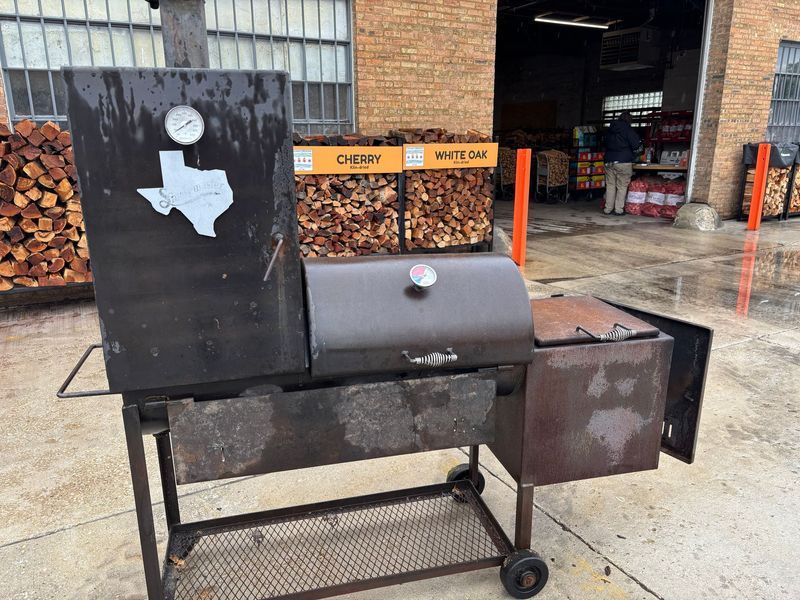 A weathered, Texas-shaped smoker with a side firebox sits outdoors near firewood.