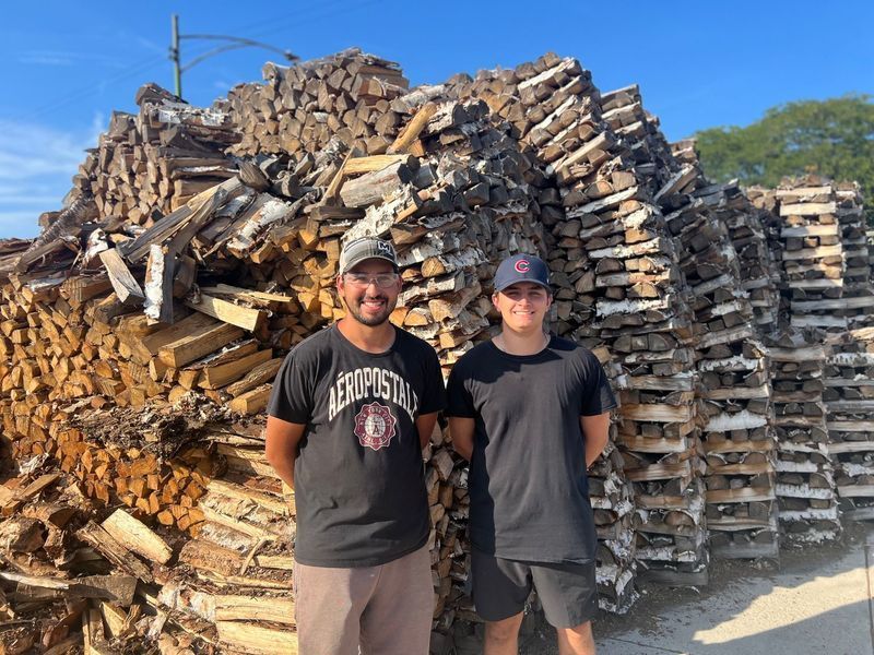 Two men stand in front of large stacks of firewood under a blue sky.