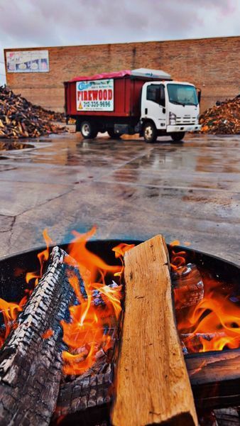Firewood truck parked near a fire pit with burning logs; brick building in background.