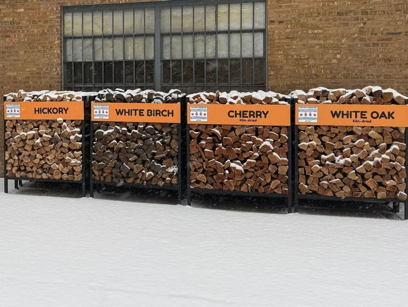 Four bins of firewood, labeled hickory, white birch, cherry, and white oak, sit outside in the snow.
