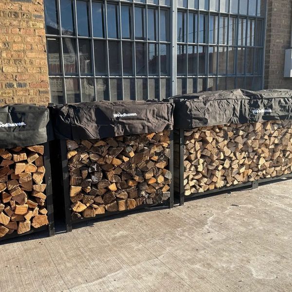 Three black metal firewood racks filled with wood, covered by black tarps, against a brick building with a window.