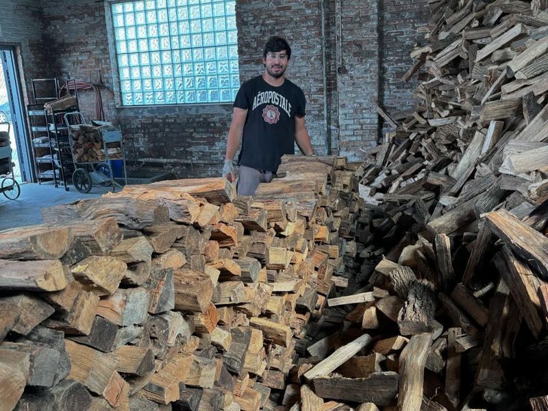 Man stands near large pile of firewood in a brick building; he wears a dark shirt and gloves.