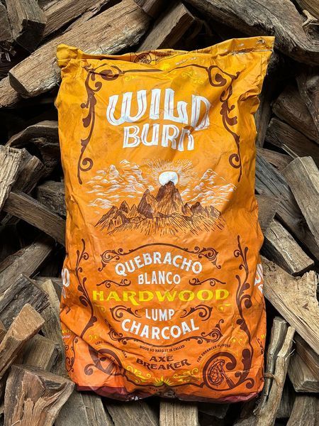 Bag of Wild Burn quebracho blanco hardwood lump charcoal against a backdrop of stacked firewood.