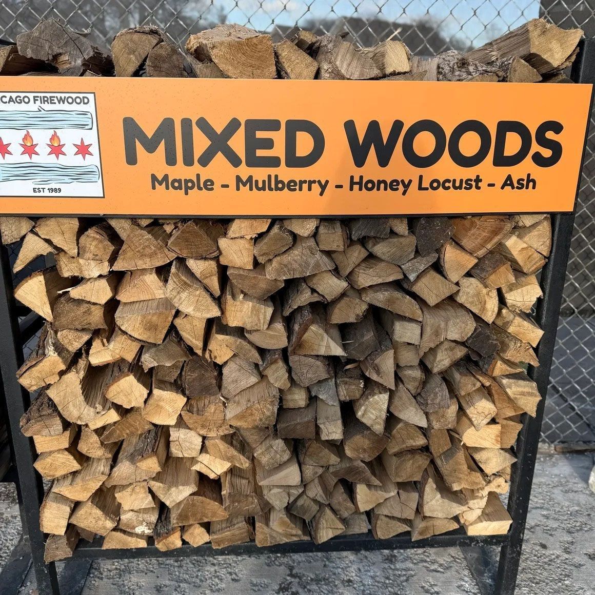 Pile of mixed firewood in a black metal frame. Sign says 