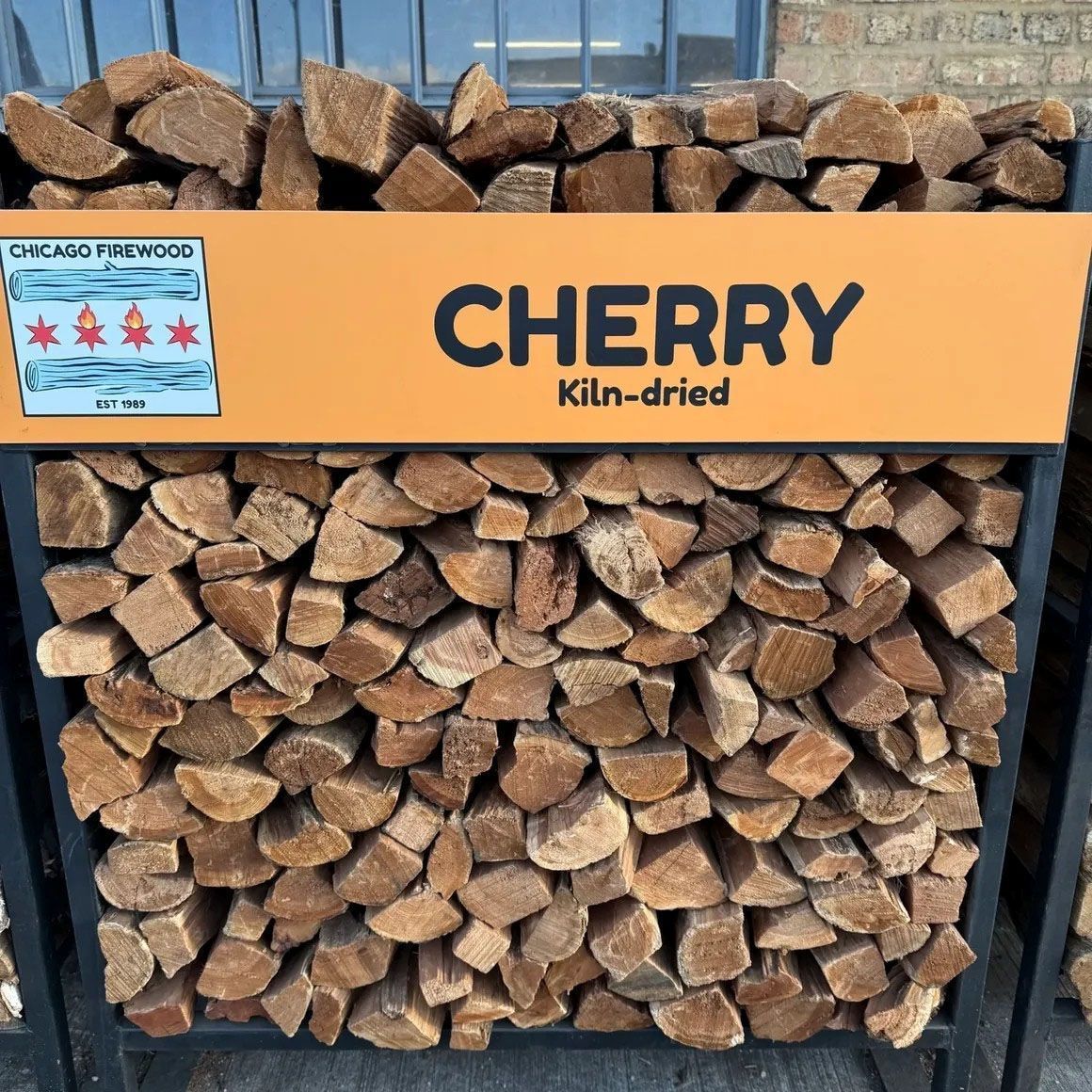 Rack of kiln-dried cherry firewood with a sign from Chicago Firewood featuring the city's flag.