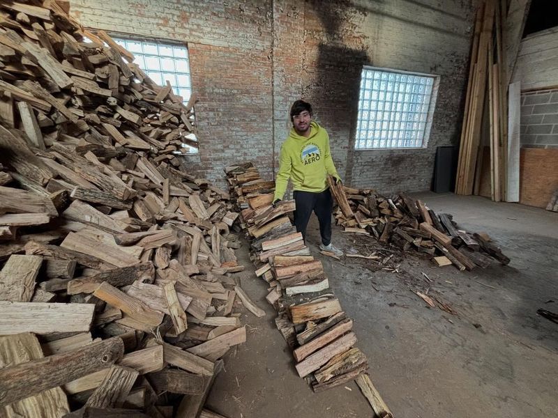 Man stacks firewood in a brick-walled warehouse. A large pile of wood stands beside him.