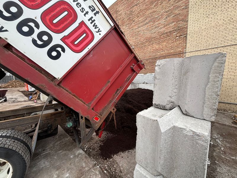 Red dump truck unloading mulch near grey retaining wall blocks.