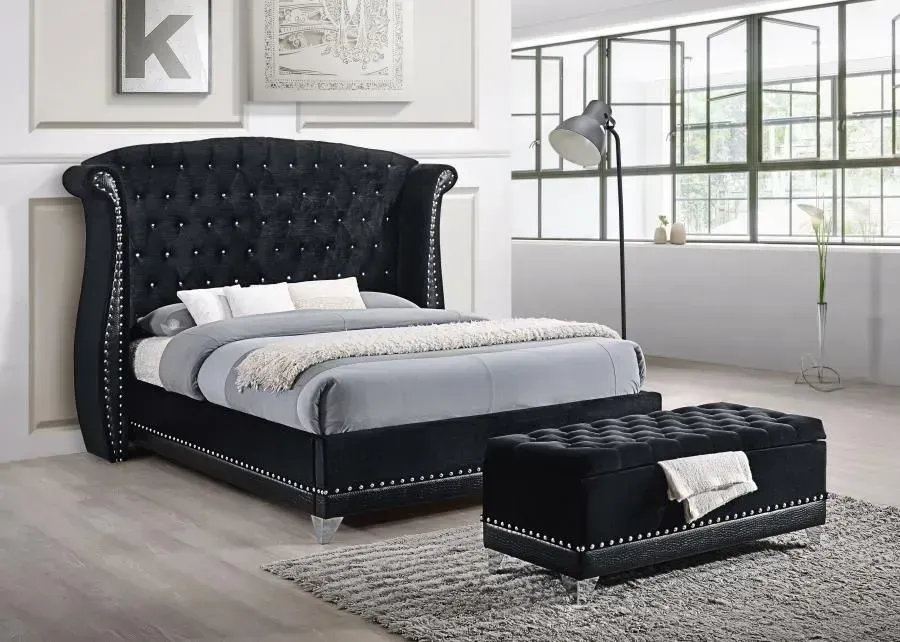 Black velvet bed with button tufting, wingback headboard, and matching ottoman on a gray rug.