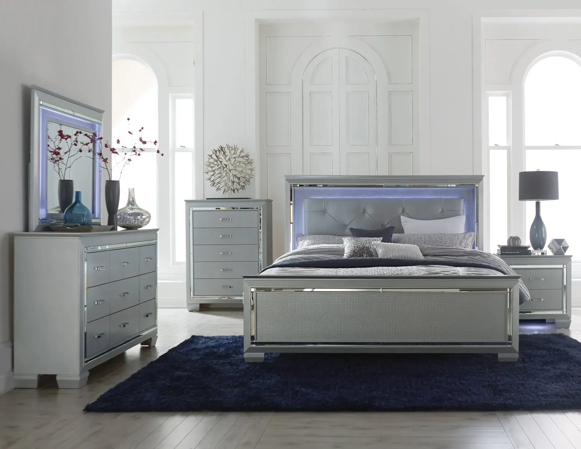 Bedroom set with mirrored accents, lit headboard, gray dresser, and a navy blue rug.