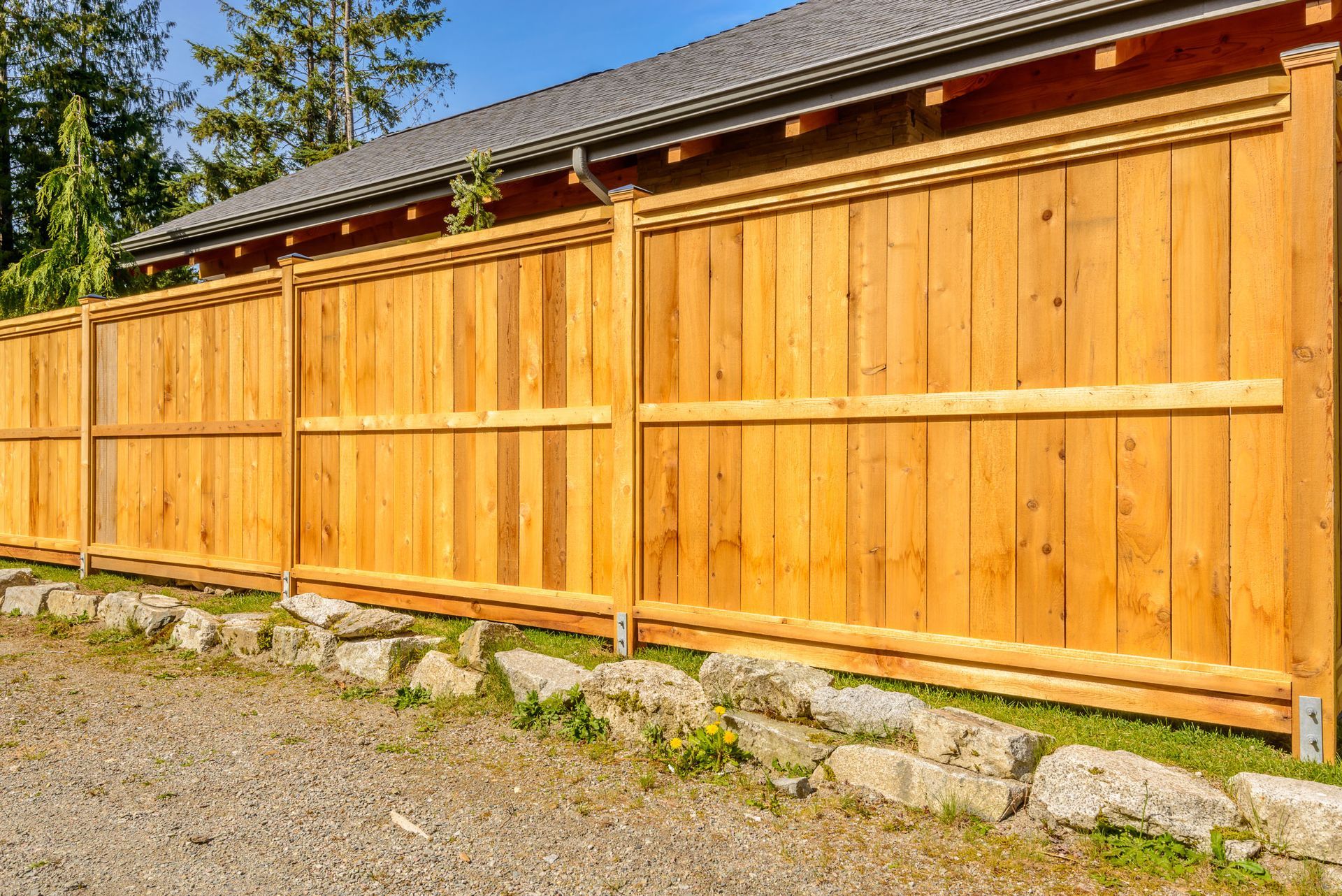 Wooden fence bordering a gravel driveway and low stone wall in front of a house.