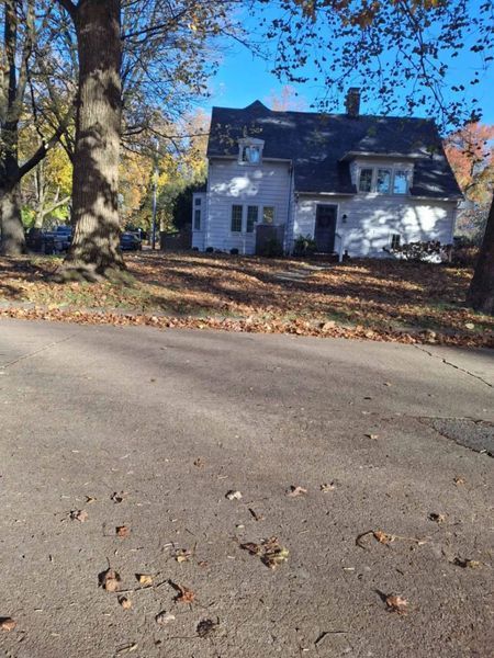 White house with dark roof on a sunny day, surrounded by trees with autumn leaves, on a street.