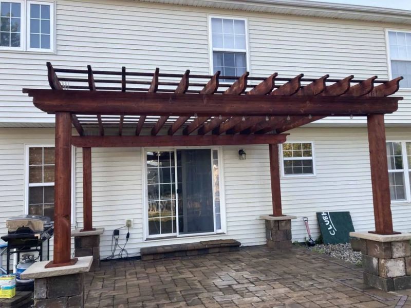 Brown pergola over a brick patio by a house with a sliding glass door.
