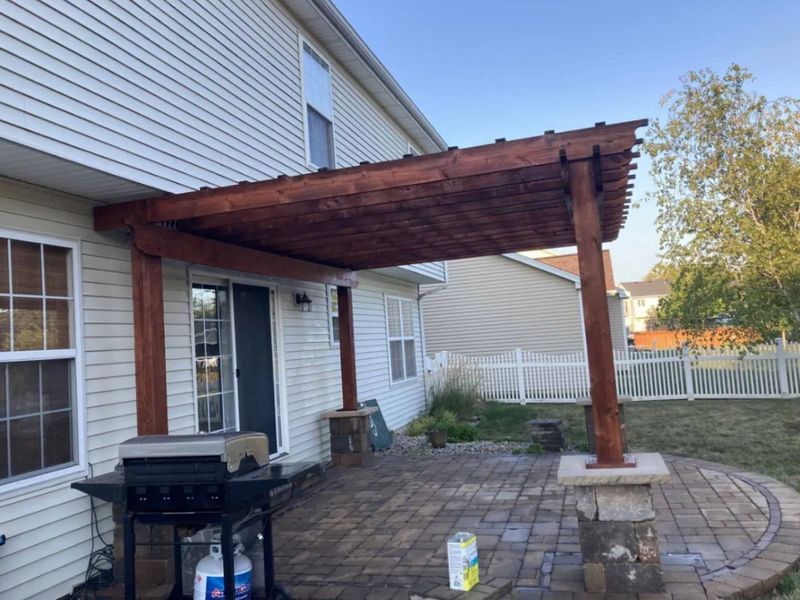Wooden pergola over a brick patio, attached to a house. Features include grill, pillars, and white fence.