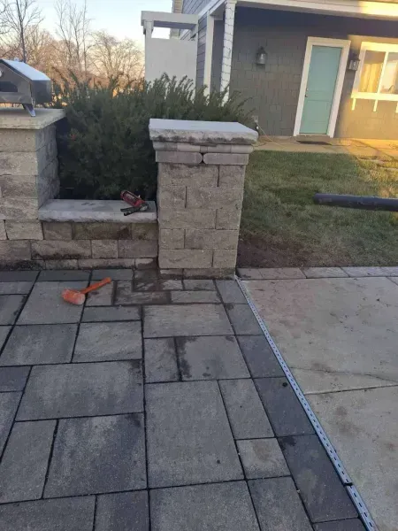 Patio with stone pavers, pillars, and a bush. Tools lie on the pavers near a pillar.
