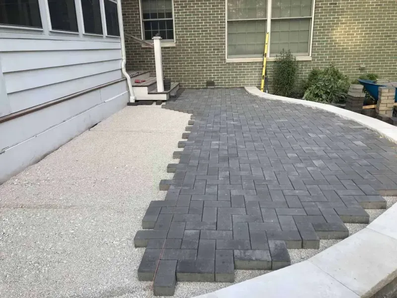 A patio being constructed with gray pavers on a gravel base next to a brick house.