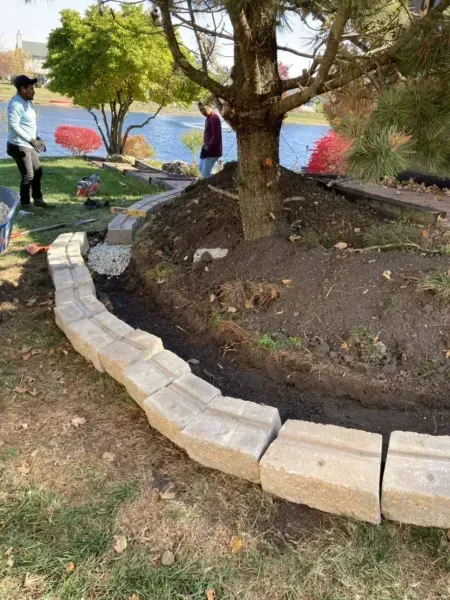 People building a stone border around a tree in a yard. Lake in background.