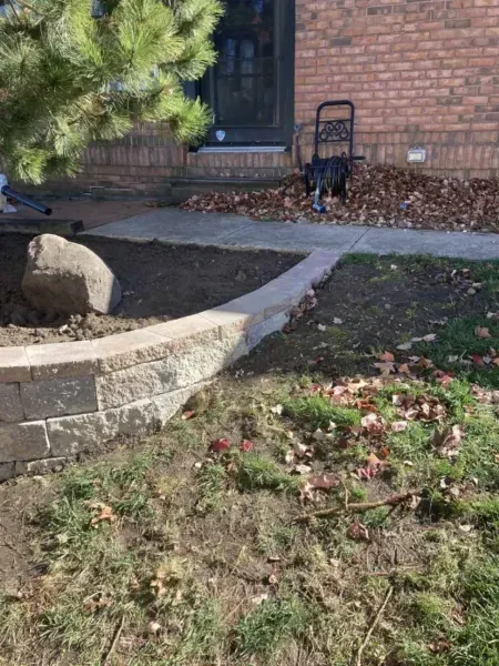 Stone retaining wall borders a lawn, with a concrete pathway leading to a house with a brick facade.
