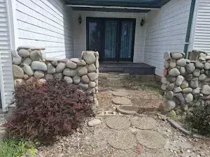 Stone pathway leads to a doorway with black doors, framed by stone walls and shrubs.