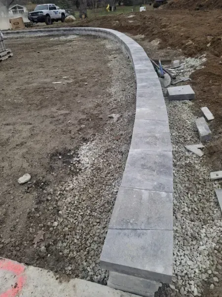 Curved stone border being constructed, gray pavers on gravel, dirt ground.