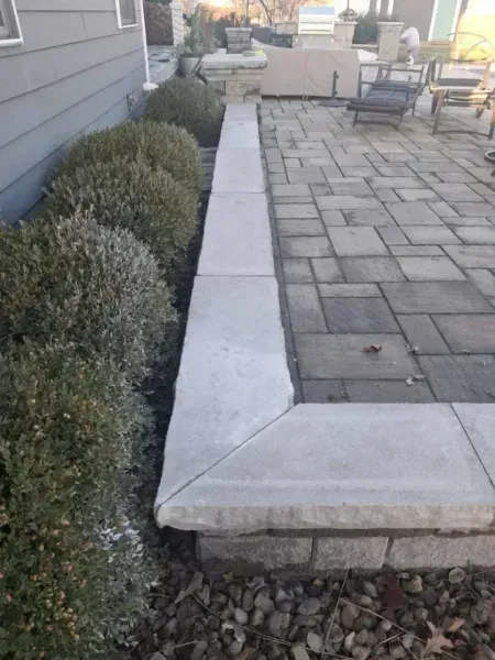 Stone patio with a low stone wall next to trimmed bushes and a house.