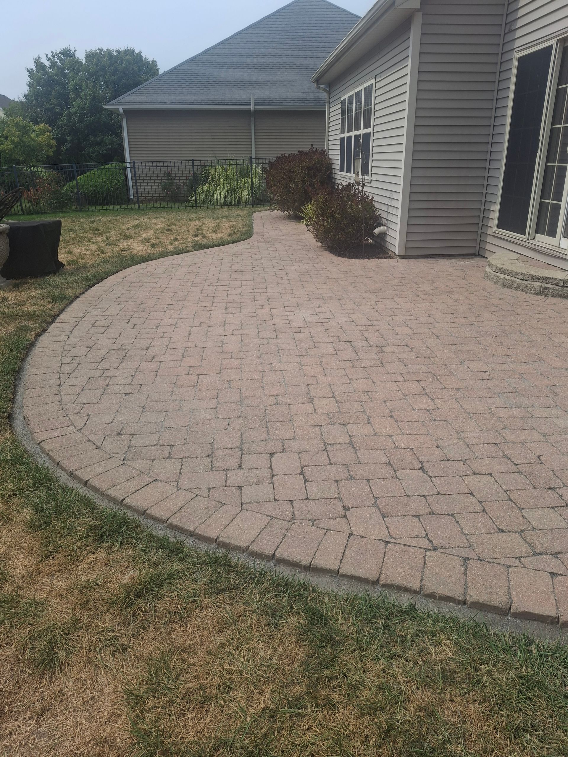 Brick patio curves near a beige house with a low roof, surrounded by patchy grass.