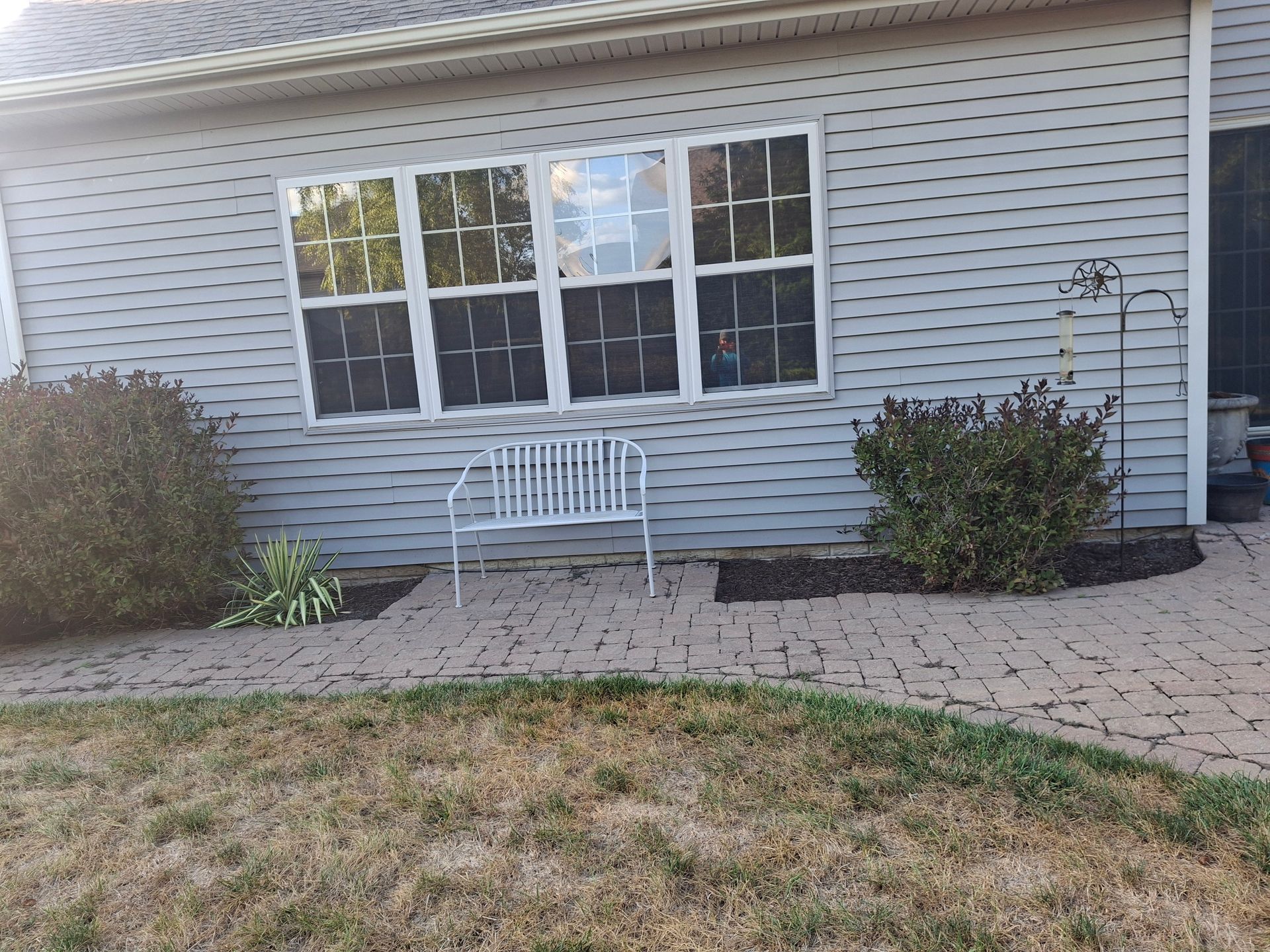 Gray house with white-framed windows, a bench, and bushes on a brick pathway.
