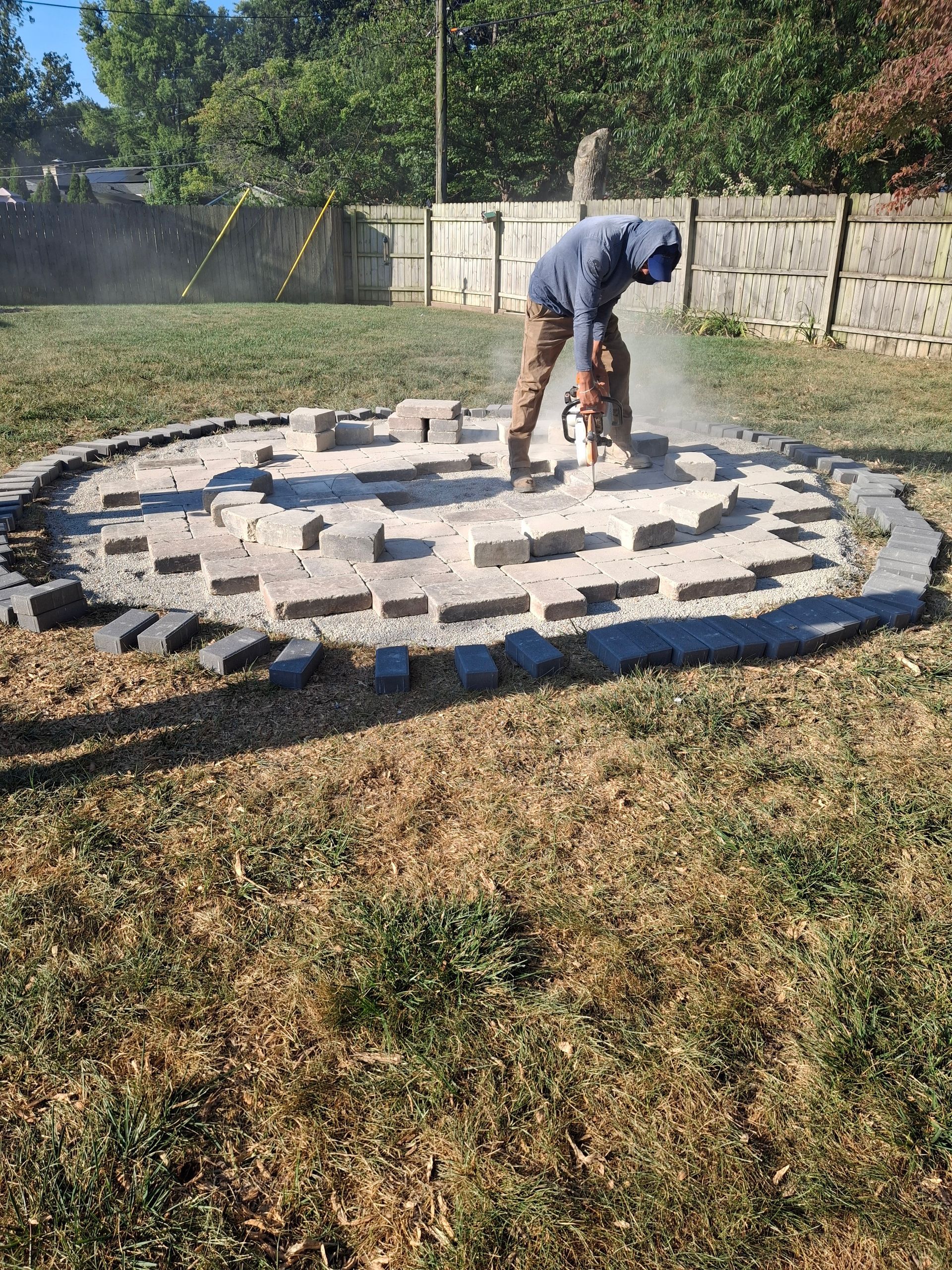 Person using jackhammer in a circular stone structure, surrounded by grass, with a fence in the background.