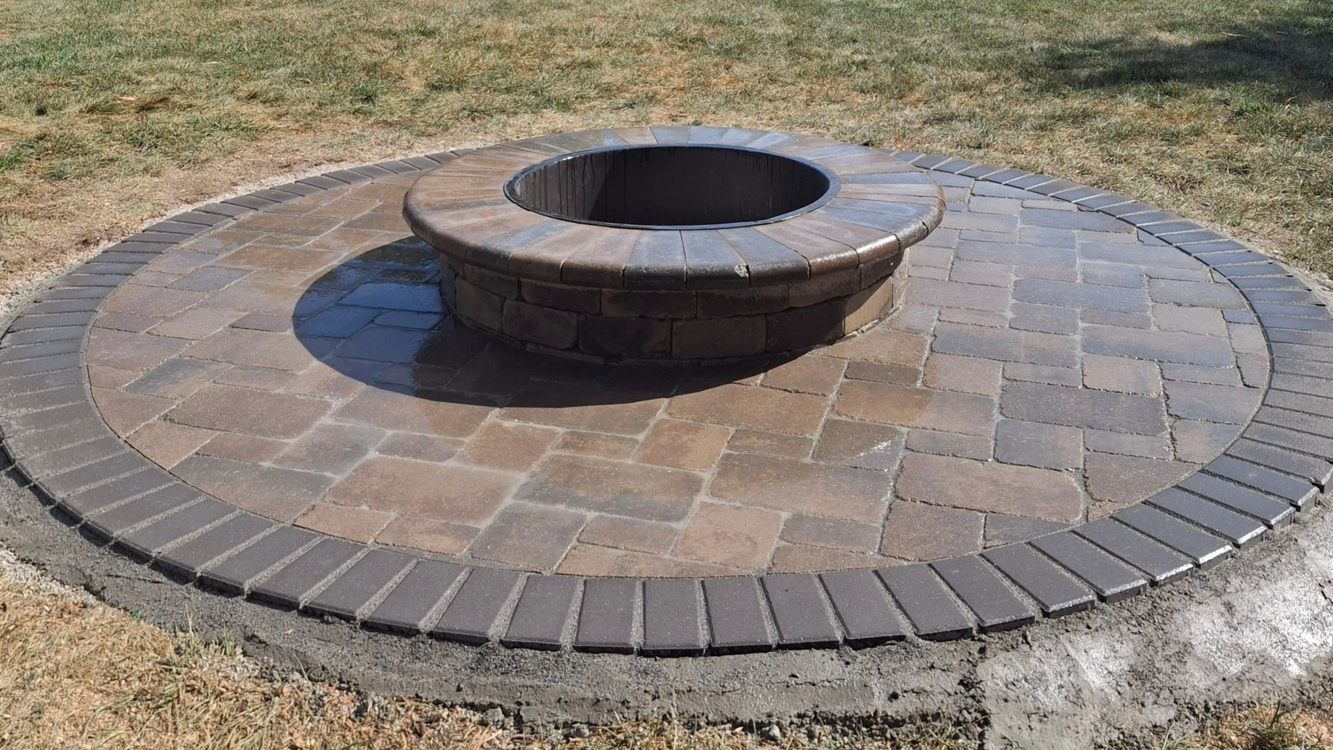 Circular stone fire pit in a backyard, surrounded by pavers and dry grass, under a blue sky.