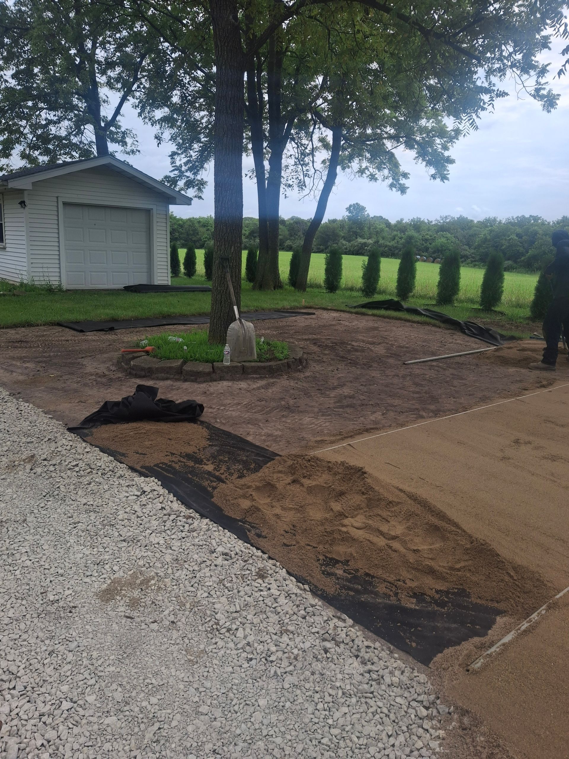Construction site with black fabric, gravel, and dirt around a tree near a white garage.