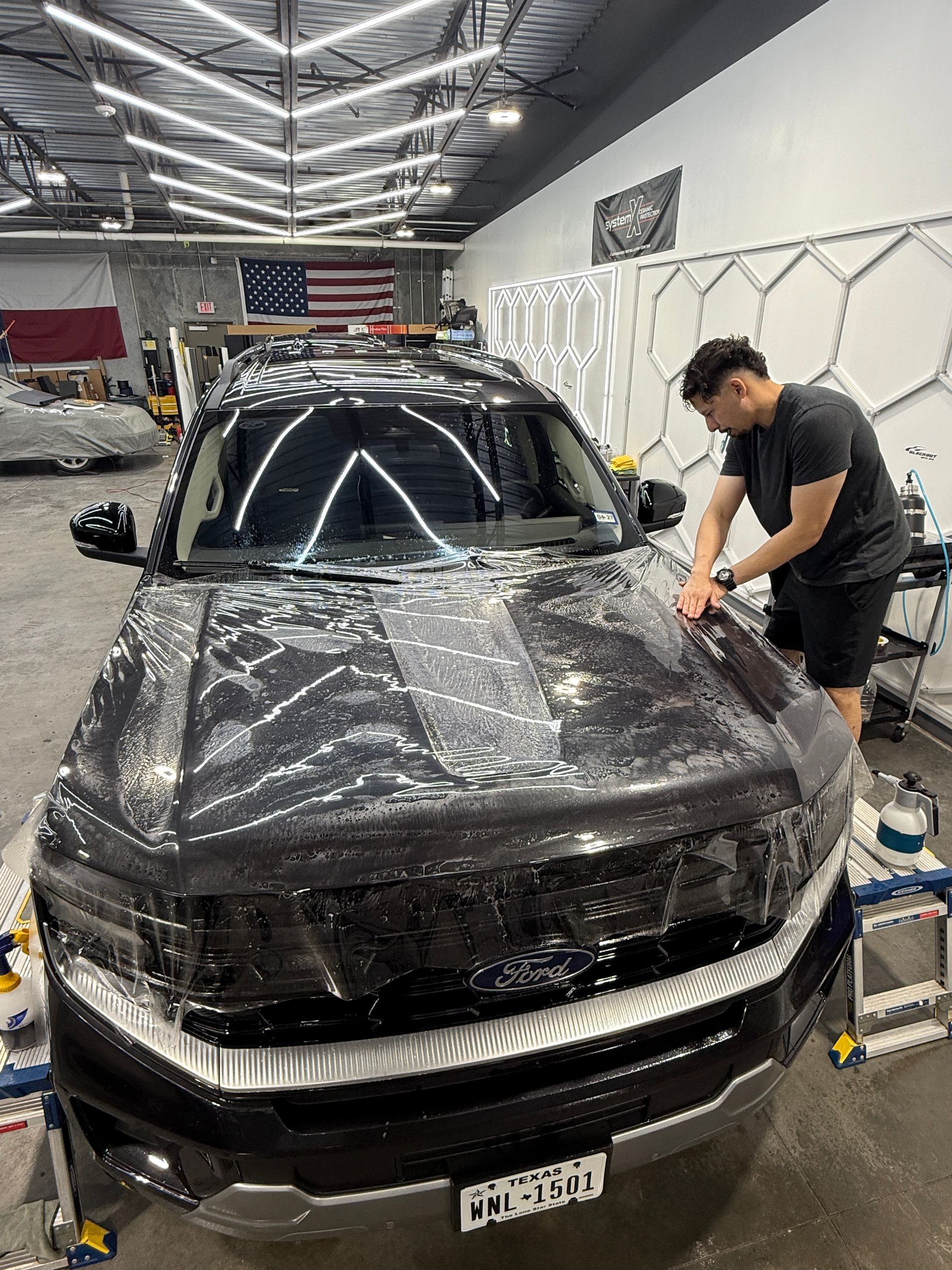 Man applying film to a black car in a well-lit shop. American flag and tools visible.