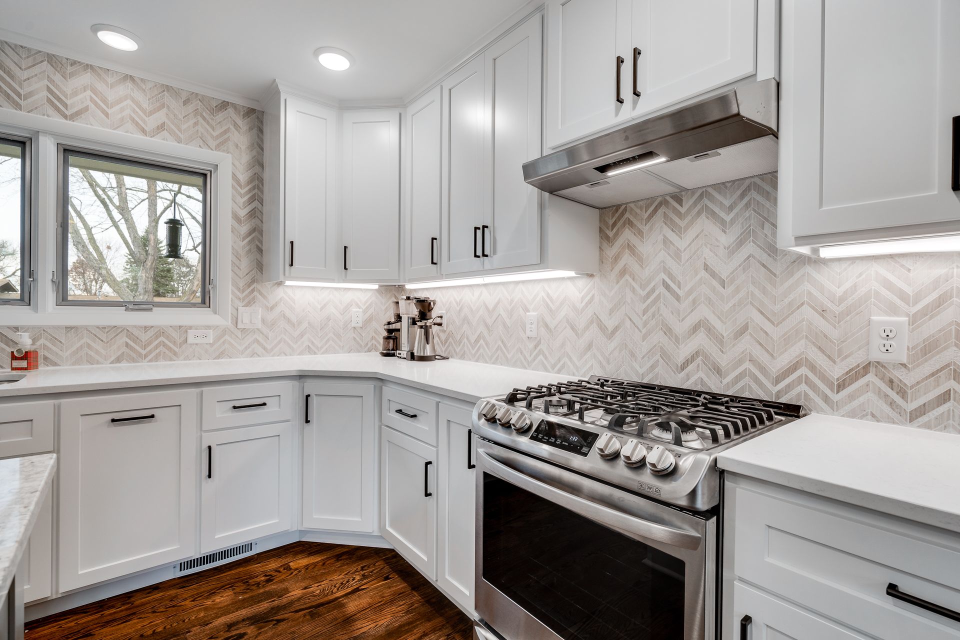 Modern white kitchen with stainless steel appliances, herringbone backsplash, and wood floors.