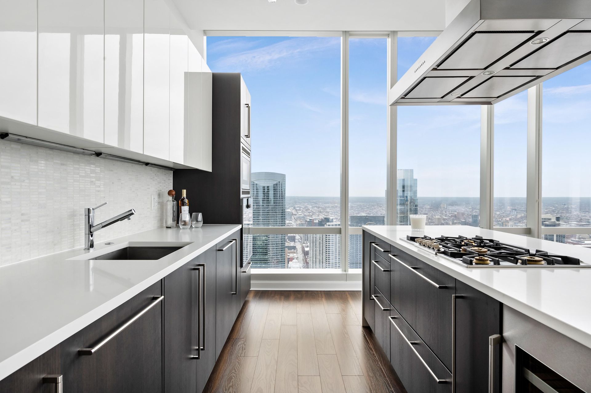 Modern kitchen with a city view through large windows; white and dark cabinets, island with stovetop.