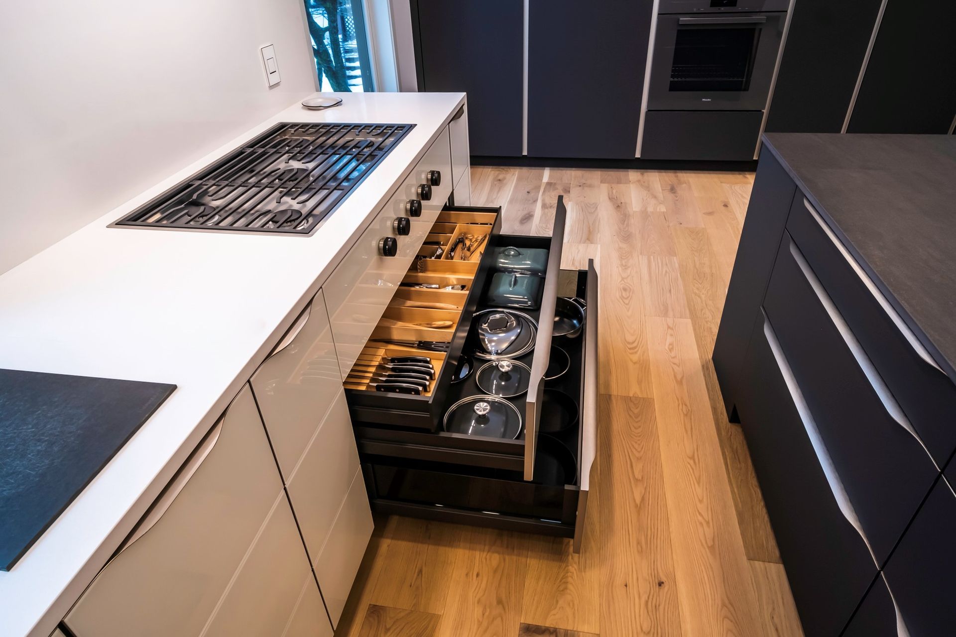 Modern kitchen with open drawer holding utensils, pots, and lids. Wooden floor, white countertops, dark cabinets.